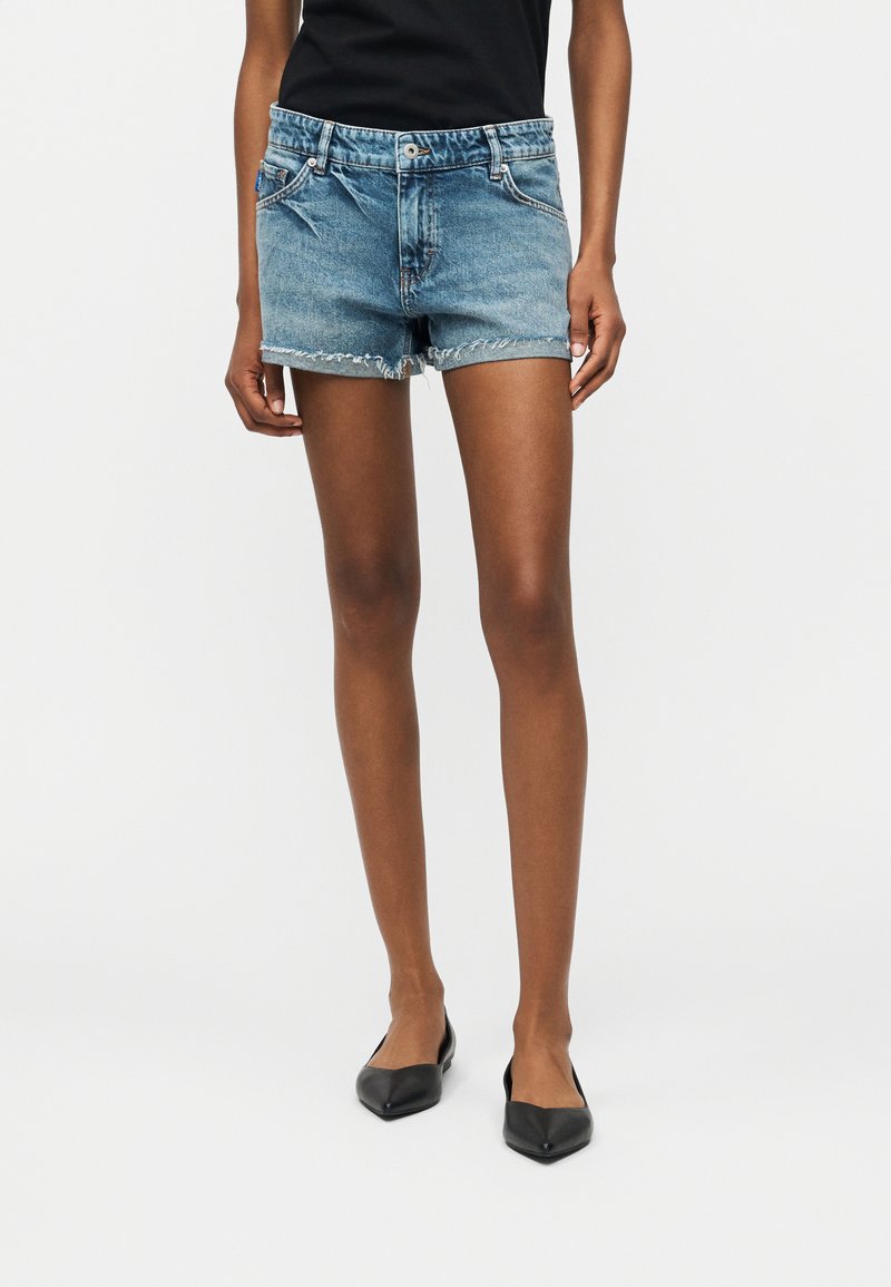 Άτομο με λεπτά πόδια που φοράει μπλε τζιν κοντά σορτς με ξεφτισμένα τελειώματα, μαύρα μυτερά παπούτσια και μαύρο τοπ μπροστά σε απλό φόντο.