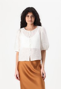 Blusa bianca con texture, maniche a sbuffo e scollatura rotonda, caratterizzata da un design frontale con bottoni. Il modello la indossa sopra una gonna marrone in satin.