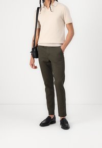 Beige polo tričko s krátkým rukávem a v-výstřihem, v kombinaci s olivovými slim-fit kalhotami. Černé mokasíny a malá černá crossbody kabelka dotvářejí celkový outfit.