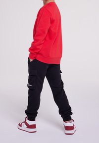Bambino che indossa una felpa rossa, pantaloni cargo neri e scarpe da ginnastica bianche e rosse, in piedi con le mani in tasca su uno sfondo neutro.