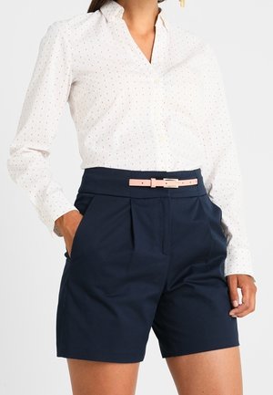 Frau trägt ein weißes, langärmeliges Hemd mit kleinen Punkten, marineblaue, hochtaillierte Shorts und einen hellrosa Gürtel, die Hände in den Taschen.