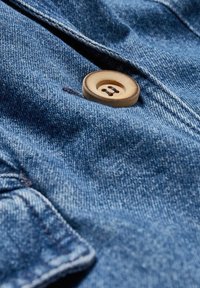 Detailní záběr hnědého čtyřdírkového knoflíku přišitého na modrý denimový materiál s viditelným prošíváním a texturou.