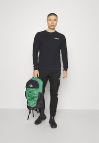Camicia nera a maniche lunghe con logo, pantaloni neri e uno zaino verde e nero con design a rete e spallacci regolabili.