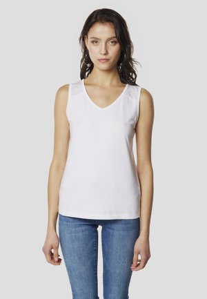 Bela majica brez rokavov z V-izrezom in teksturiranimi poudarki na ramenih, v kombinaciji s prilegajočimi se modrimi kavbojkami. Gladek material, priložnostna zasnova.