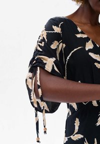 Vrouw in een zwarte jurk met een beige bloemenprint, met een V-hals en gerimpelde korte mouwen met hangende strikjes en houten kralen.