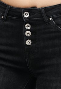 Μαύρο τζιν από denim με τέσσερα ορατά ασημένια κουμπιά που κλείνουν το μπροστινό μέρος και μερική όψη στη μέση και στα θηλύκια της ζώνης.