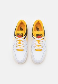 Vita sneakers med gula accenter, svart häl och vita snören. Har en basketgrafik och texten "FORCE" på tungan. Slät yta.
