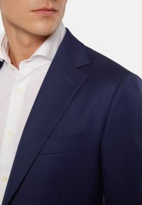 Námořnická modrá sako s texturovanou látkou, jednou náprsní kapsou a špičatými klopami. Pod ním je bílá košile s límcem a žlutými knoflíky.