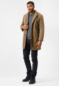 Tan vævet frakke med revers og to lommer, båret over en grå sweater og sorte bukser. Sorte snørebåndsstøvler fuldender outfittet.