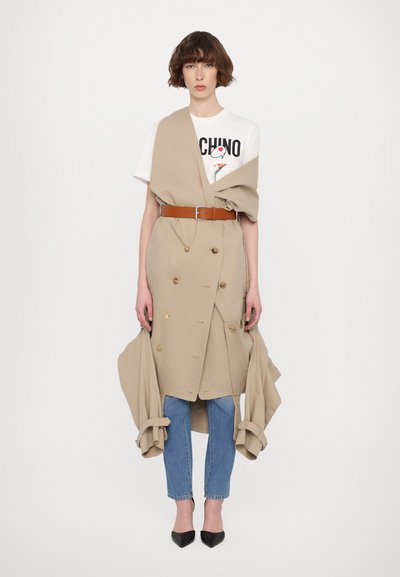 Beige, doppelt geknöpfter Trenchcoat mit großen Knöpfen, tailliert mit einem Gürtel, über einem weißen Grafik-T-Shirt und hellblauen Jeans getragen. Schwarze, spitze High Heels.