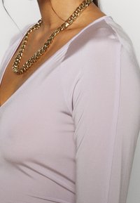 Lilafarbenes Langarmoberteil mit tiefem V-Ausschnitt, aus einem glatten Stoff gefertigt, ergänzt durch eine große goldene Kettenhalskette.