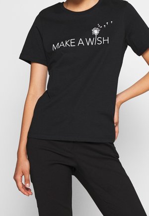 Person iført en sort t-shirt med hvid tekst "MAKE A WISH" og et mælkebøttefrø, der blæser grafikken på brystet.
