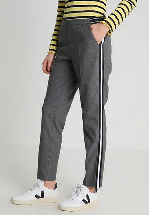 Benen met een grijze broek met zwarte en witte zijkstrepen, witte sneakers met zwarte V-logo's, en een gele top met lange mouwen met zwarte strepen.