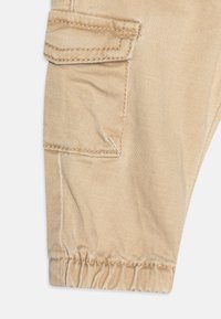 Beige cargobroek heeft een zijzak, elastische boorden en fijne stiksels. De stof heeft een gladde textuur met een lichte glans.
