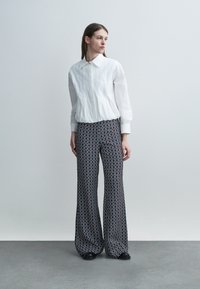 Donna in piedi, indossa una blusa bianca a maniche lunghe e pantaloni a vita alta e gamba larga con un motivo circolare nero e bianco.