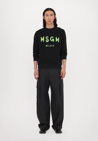 Zwarte sweatshirt met groene tekst "MSGM MILANO", gecombineerd met losse zwarte cargobroek. De stof lijkt soepel met lange mouwen.