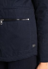 Marineblå jakke med en frontlomme med lynlås og et logo-tag. Stoffet ser glat ud med synlige syningsdetaljer og en struktureret form.