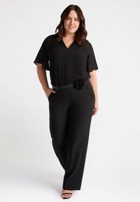 Blusa nera con maniche corte e dettagli plissettati, abbinata a pantaloni neri sartoriali. Presenta una cintura nera con un accento floreale.