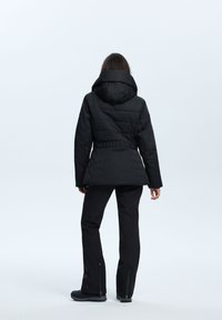 Schwarze gesteppte Jacke mit Kapuze, tailliertem Bund und langen Ärmeln; kombiniert mit schwarzen Hosen mit Reißverschlussabschlüssen und einfarbigem Design.
