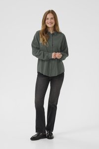 Žena koja se smije stoji s rukama prekriženim, noseći zelenu košulju s gumbima, izblijedjele crne traperice i crne mokasinke na bijeloj pozadini.