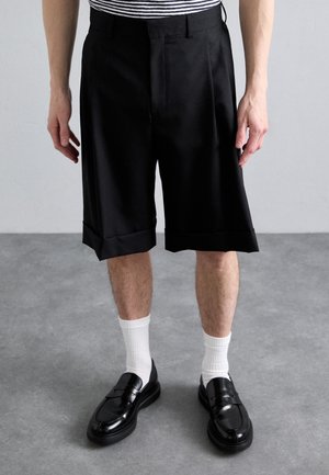 Pantalones cortos negros de corte entallado con pliegues, hasta la rodilla, que cuentan con un amplio dobladillo. Combinados con calcetines blancos y zapatos de charol negros. Camisa a rayas visible.