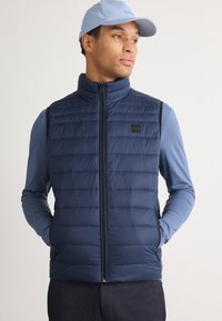 Tums fuscia krāsas izolēts veste ar vertikālu quilted strukt�ūru, ar rāvējslēdzēja aizdari un sānu kabatām; uzvilkta virs gaiši zilas garām piedurknēm krekla.