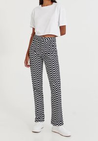 Persona vestida con pantalones de patrón ondulado en blanco y negro, una camiseta blanca cropped sencilla y zapatillas blancas, de pie frente a un fondo liso.