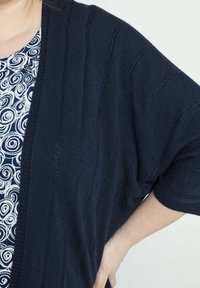 Mörkblå stickad cardigan med ribbad textur och vida ärmar, lager-på-lager över en vit och mörkblå top med swirl-mönster.