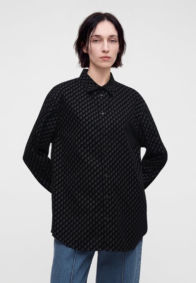 Jeune personne aux cheveux courts et foncés portant une chemise noire à motifs à manches longues et un jean bleu, debout devant un fond uni.