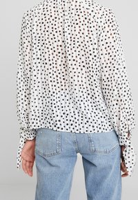 Blusa bianca con pois neri e marroni, dotata di maniche lunghe e polsini annodati, abbinata a jeans azzurri chiari. Tessuto testurizzato e semi-trasparente.