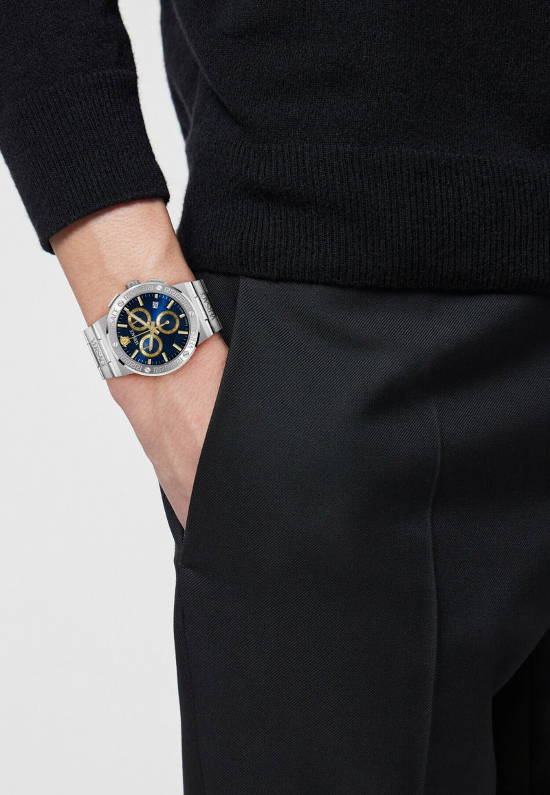 Silver stainless steel watch with a blue dial, golden accents, and chronograph features, paired with a black knit sweater and dark pants.