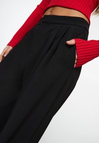 Osoba v černých kalhotách s vysokým pasem a sklady, v zkrácené červené žebrované svetru, s jednou rukou schovanou v kapse kalhot.