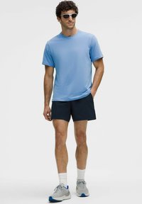 Lichtblauw t-shirt met korte mouwen, gecombineerd met donkere shorts, grijze sportschoenen en witte sokken. Geaccessoired met witte zonnebril.