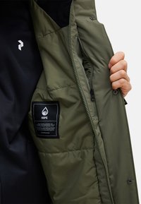 Green insulated jacket interior with zippered pocket and care label. Features black lining and logo on chest. Smooth, textured fabric.