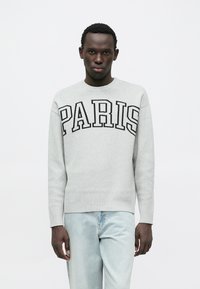 Sweat-shirt gris avec l'inscription noire "PARIS" sur la poitrine, col rond et poignets côtelés, porté avec un jean bleu clair.