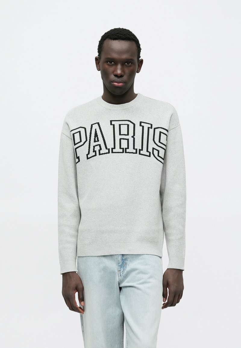 Sweat-shirt gris avec l'inscription noire "PARIS" sur la poitrine, col rond et poignets côtelés, porté avec un jean bleu clair.