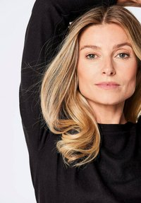 Porträt einer Person mit langen, welligen blonden Haaren, die ein schwarzes Oberteil trägt. Der Hintergrund ist weiß und hebt die Gesichtszüge sowie die Haarstruktur hervor.