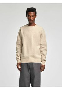 Beżowy wełniany sweter z okrągłym dekoltem i ściągaczami przy mankietach, zestawiony ze szarymi spodniami w prążki. Tekstura wydaje się miękka i lekko puszysta.