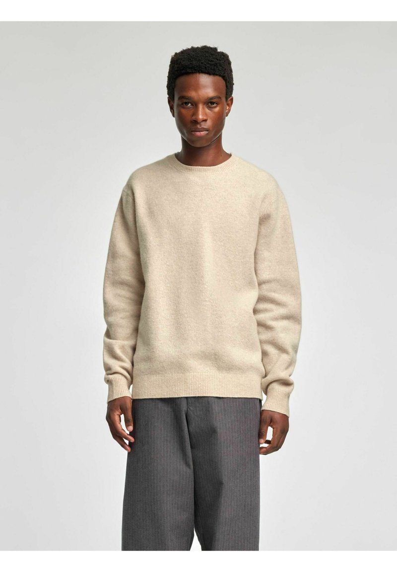 Beżowy wełniany sweter z okrągłym dekoltem i ściągaczami przy mankietach, zestawiony ze szarymi spodniami w prążki. Tekstura wydaje się miękka i lekko puszysta.