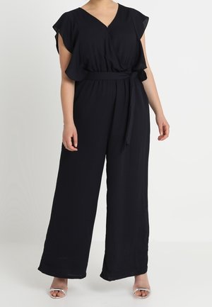 Vrouw draagt een zwarte jumpsuit met wijd uitlopende pijpen, zwierige mouwen, een V-hals en een taillekoord, gecombineerd met zilveren open teen hakken.