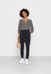 Blusa a righe in bianco e nero con dettaglio a occhio di bue, abbinata a pantaloni jogger blu scuro con tasche con zip. Sneakers bianche e borsa intrecciata.