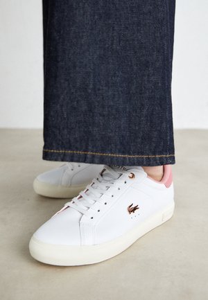 Zapatillas blancas con detalles rosas y logo de cocodrilo marrón, usadas con pantalones vaqueros azul oscuro, de pie sobre un suelo claro.