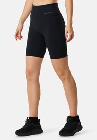 Zwarte high-waisted fietsbroek van rekbaar materiaal, met een soepele textuur en een logo op de tailleband, gecombineerd met zwarte sportschoenen.