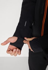 Čierna bunda s oranžovým zipsom, s textúrovanými manžetami a otvorom pre palce. Materiál vyzerá odolný s kombináciou hladkých a textúrovaných povrchov.