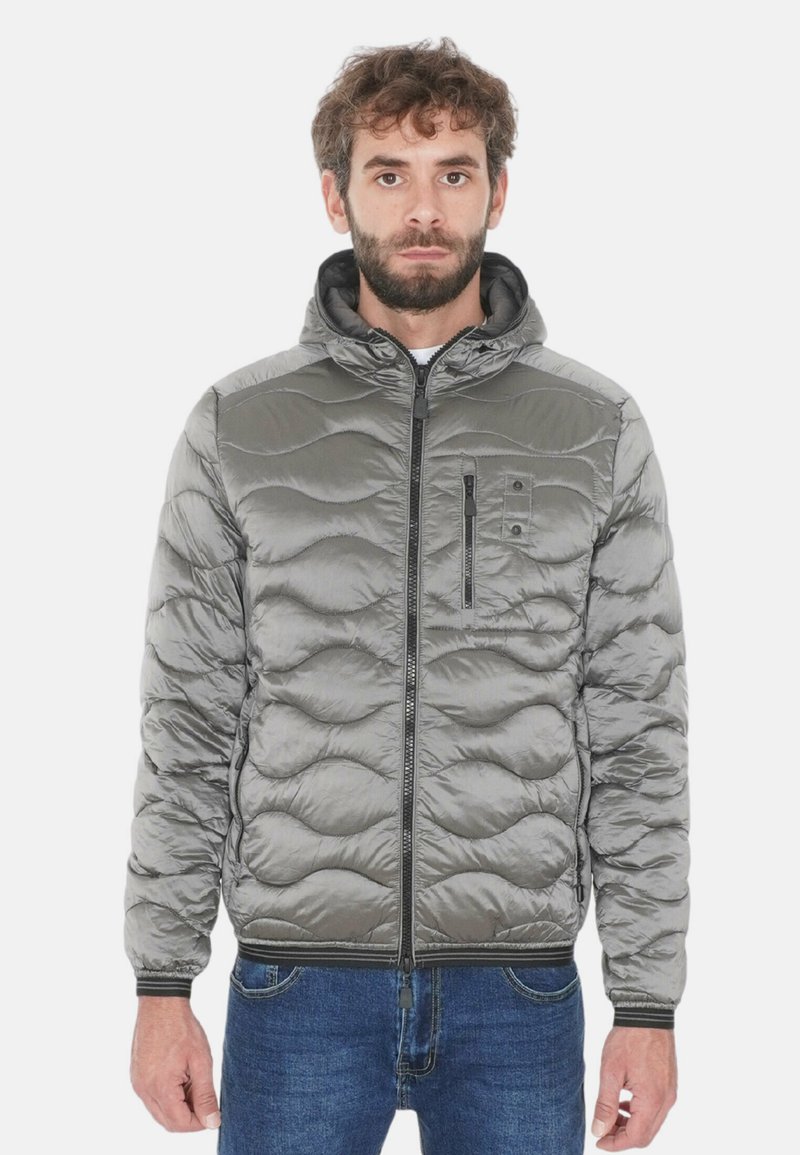 Giacca imbottita grigia con cappuccio, presenta un motivo trapuntato, chiusura con zip e una tasca sul petto. Indossata con jeans di jeans blu.