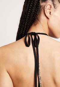 Svart halterneck-top med justerbar knytning baktill; har slät tygkvalitet och en subtil lyster; tatuering synlig på övre delen av ryggen.