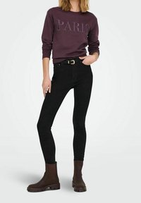 Jeans skinny neri abbinati a una felpa bordeaux con il testo "PARIGI". Il modello indossa stivaletti marroni con una suola spessa.