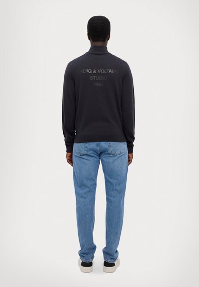 Schwarzer Strickpullover mit hohem Kragen, versehen mit eingeprägtem Logotext auf der Rückseite. Getragen mit hellblauen Jeans und schwarzen Turnschuhen.