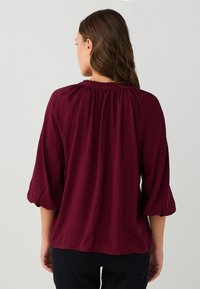 Frau mit langen braunen Haaren, die eine burgunderfarbene Bluse mit gerafftem Ausschnitt und dreiviertel langen Puffärmeln trägt, von hinten blickend, vor einem schlichten Hintergrund.