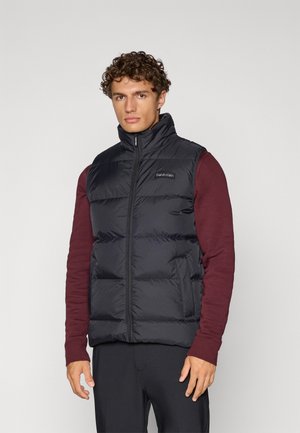 Juodas pūkinis liemenė su aukštu apykakle, turi užtrauktuką ir Calvin Klein logotipą. Dėvima per bordo ilgomis rankovėmis marškinėlius, lengvas dizainas.